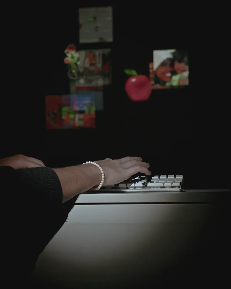 Hands typing on a lit keyboard in a dark room, images projected on the wall behind