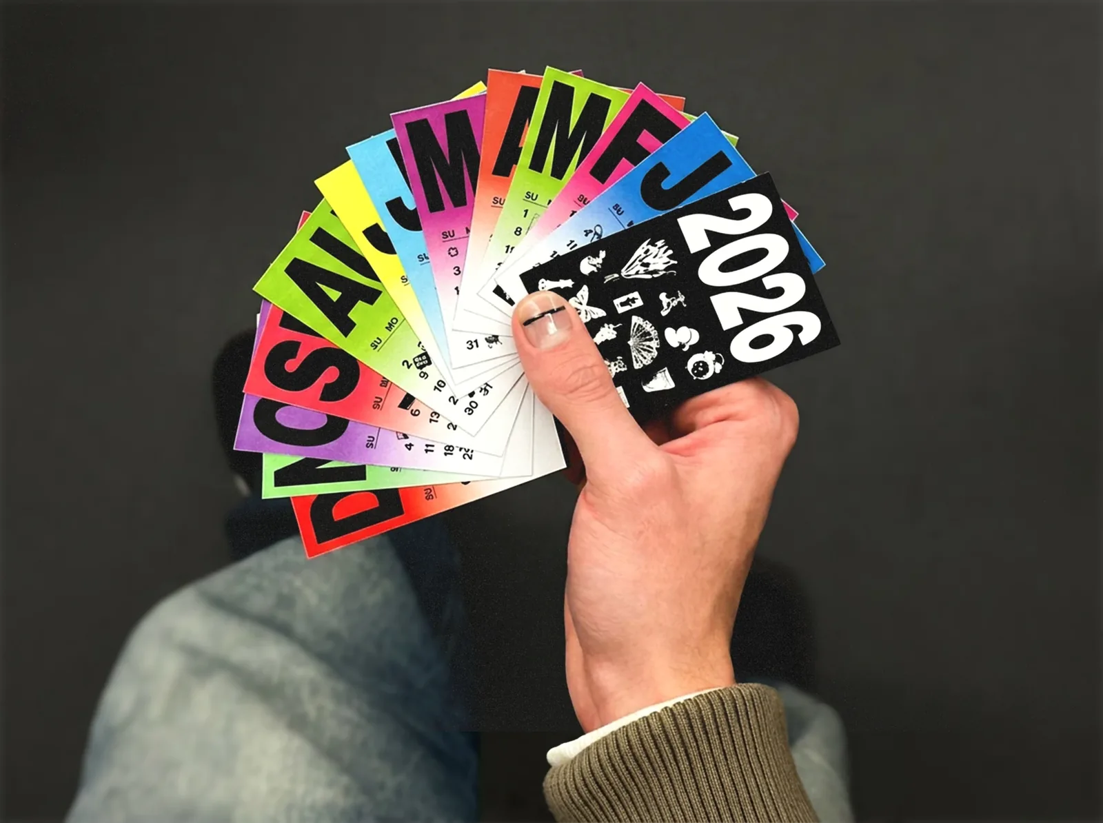 A hand fanning out the twelve colourful month cards against a dark background